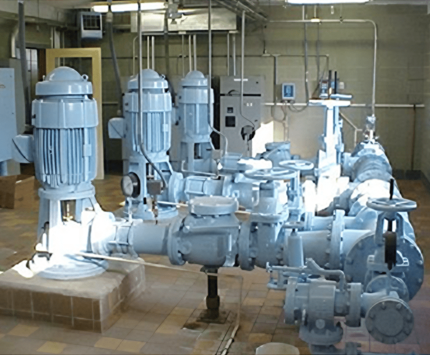 An industrial pump room featuring three large vertical electric motors in light blue, mounted above horizontal centrifugal pumps with an intricate network of piping and control systems in a municipal or water treatment facility