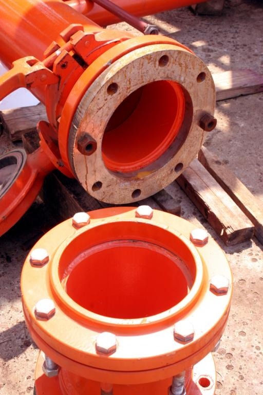 Close-up of bright orange fire hydrant flanges with bolted connections, showing the pipe coupling system with visible bolt holes and weathered interior surfaces at an outdoor construction or maintenance site