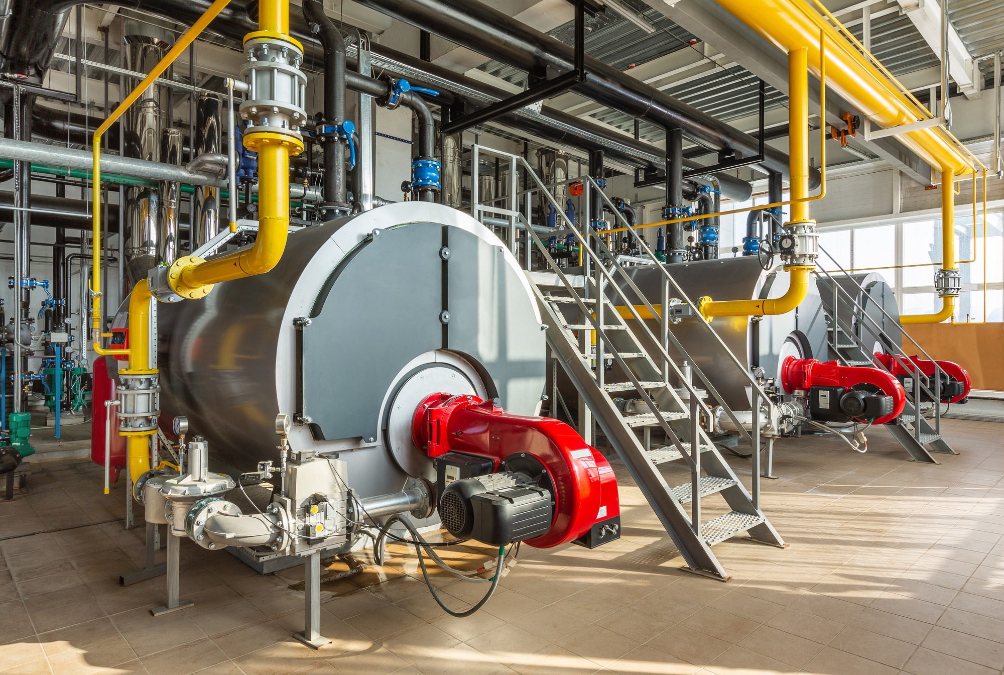 The interior of an industrial boiler room with three large boilers, many pipes, valves