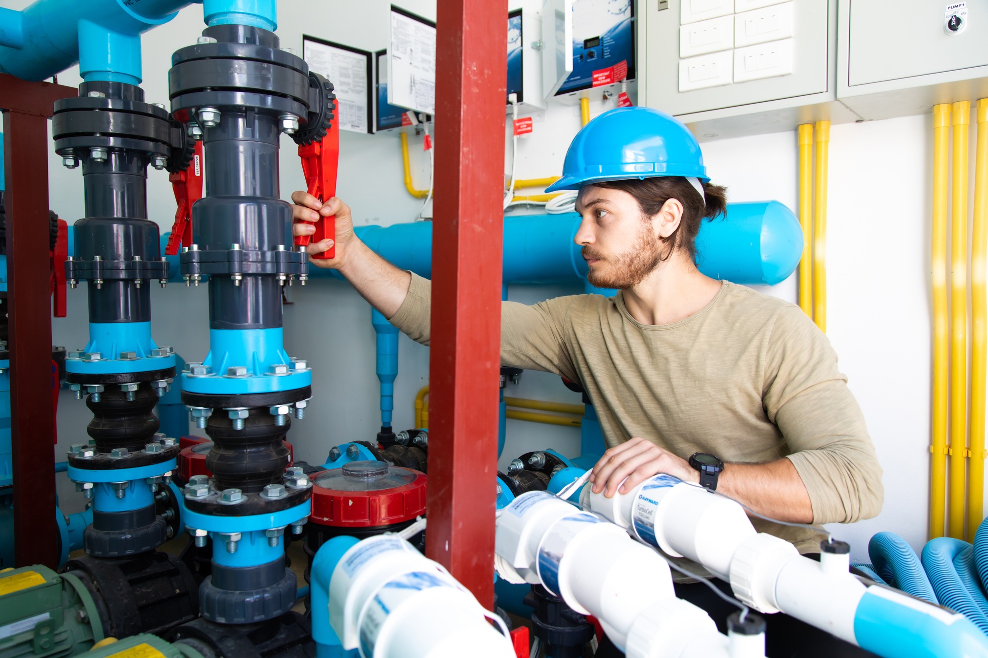 A technician wearing a blue hard hat adjusts a red valve on industrial piping equipment, surrounded by bright blue and gray flanged connections, white PVC pipes, and yellow conduit in a well-organized mechanical room